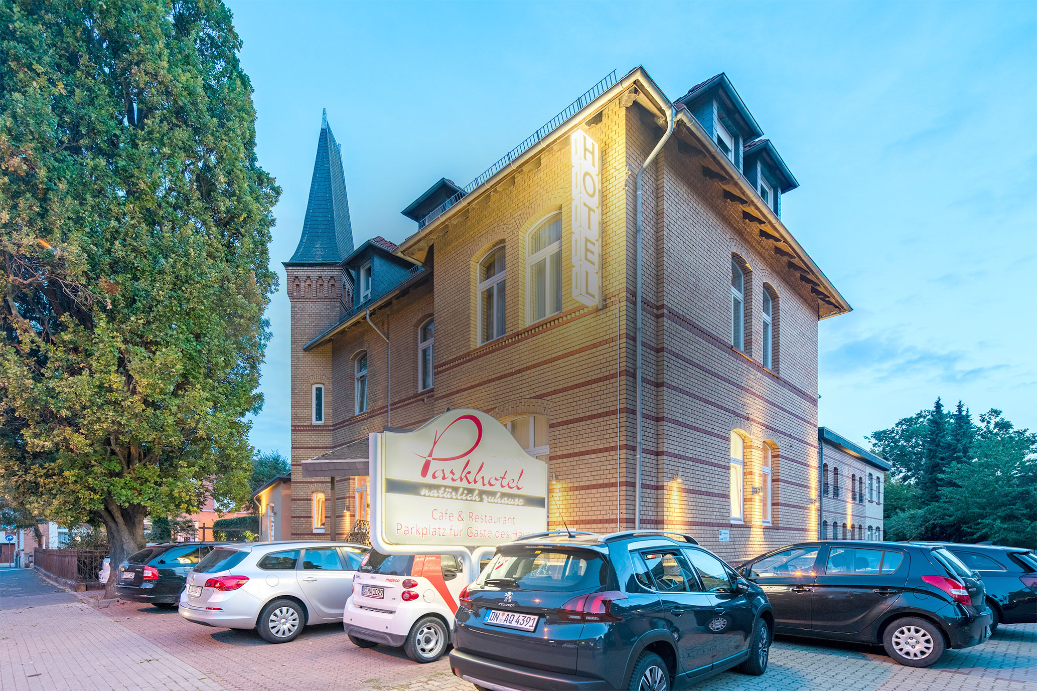 Das Parkhotel mit gelber Backsteinfassade und Turm, beleuchteter Hoteleinschrift, Parkplatz und mehreren geparkten Autos vor dem Gebäude. Parkhotel Helmstedt