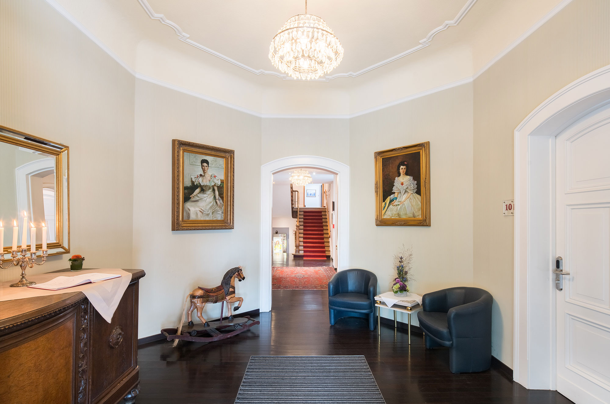 Hotelhalle mit Gemälden, Schaukelpferd und klassischem Mobiliar. Im Hintergrund Treppe mit rotem Teppich und Kronleuchter. Parkhotel Helmstedt